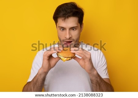 Guilty Pleasure Concept. Portrait of hungry funny young man holding burger in hands and eating unhealthy fastfood, excited starving guy wants sandwish licking his lips, yellow orange studio wall Royalty-Free Stock Photo #2138088323