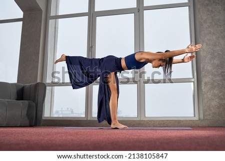 Side view of sporty girl practicing yoga in palazzo pants indoors. Flexible brunette woman standing on one leg with hands straight, stretching. Concept of yoga and new age,