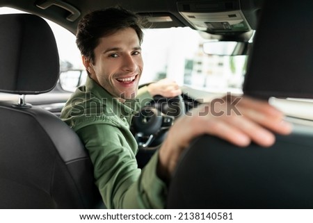 Cheerful young man driving new automobile after purchase, sitting in driver's seat of car at dealership. Happy millennial guy looking back and smiling, buying modern auto at showroom Royalty-Free Stock Photo #2138140581