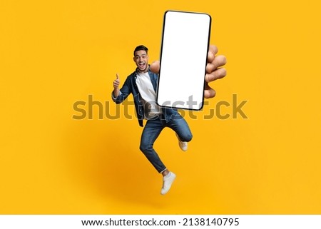 Cheerful handsome millennial middle eastern guy in styish outfit jumping up with modern smartphone, showing white empty screen and thumb up, recommending online offer, yellow background, copy space