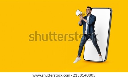 Handsome young arab man jumping out of huge smartphone with white empty screen, holding megaphone over yellow studio background, making important announcement, panorama with copy space Royalty-Free Stock Photo #2138140805