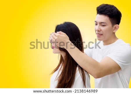 Young man closing eyes of his girlfriend while making a surprise in the studio with yellow background