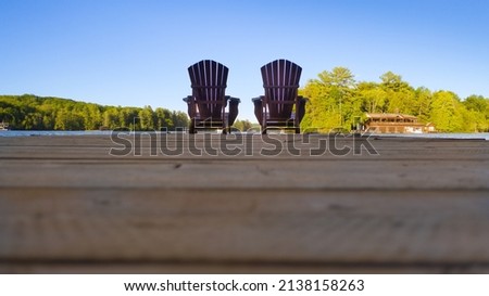 Two empty Adirondack chairs sitting on a dock on a lake in Muskoka, Ontario Canada. Calm morning on a cottege pier. Royalty-Free Stock Photo #2138158263