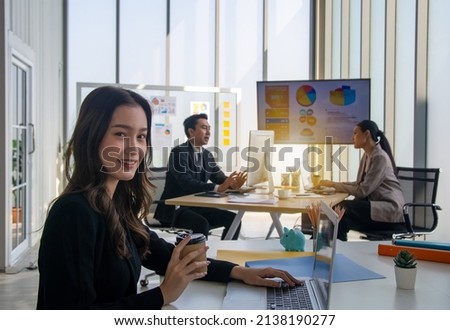 Cozy work environment. Successful smiling businesswoman sitting at the table working on notebook and holding coffee cup on the background of defocus two business people.