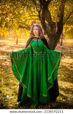A beautiful girl in a medieval green dress with golden braid. Queen in a cloak and a blio dress in the autumn forest. historical image Royalty-Free Stock Photo #2138192211
