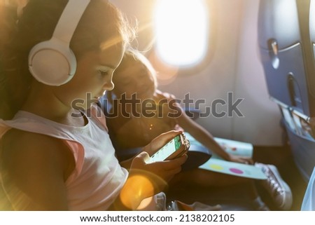 Kid schoolgirl in headphones watches cartoons in a phone on an airplane. little girl travels in an airplane, sitting in her seat, playing with a tablet computer, phone and headphones