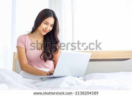 Asian young happy beautiful female model in casual pajama outfit sitting smiling under white clean blanket on bed in bedroom browsing surfing internet shopping online via laptop notebook computer.