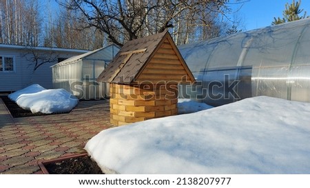 Early spring at the dacha. View of a closed well in the form of a small wooden house and a greenhouse. Garden paths without snow, snowdrifts on flower beds and beds