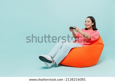 Full body young smiling happy woman of Asian ethnicity 20s wearing pink sweater sit in bag chair hold in hand play pc game with joystick console isolated on pastel plain light blue background studio