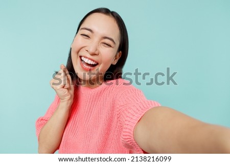 Close up young happy woman of Asian ethnicity 20s in pink sweater do selfie shot pov on mobile phone do winner gesture isolated on pastel plain light blue background studio. People lifestyle concept