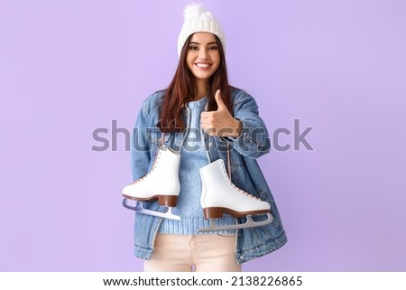 Beautiful young woman in winter clothes and with ice skates showing thumb-up gesture on color background