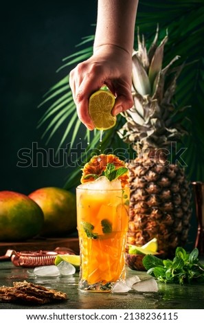 Summer cocktail with vodka, pineapple juice, mango, ice. Long drink or cold mocktail. Bartender hand squeezes lime juice, frozen motion and flying drops. Tropical background with palm leaves 