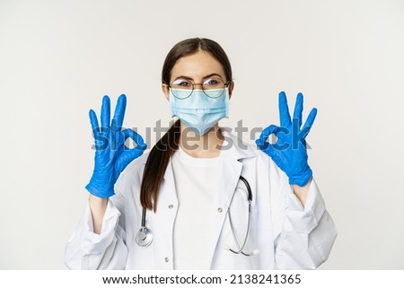 Close up portrait of woman doctor, physician in face mask from coronavirus, showing okay sign in approval, recommending, praise and like smth good, standing over white background