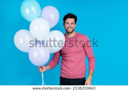 Celebration Concepts. Smiling Funny Caucasian Guy Handsome Brunet Man With Bunch of Colorful Air Balloons Posing in Pink Jumper Against Blue Background. Horizontal Image