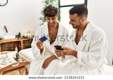 Two hispanic men couple holding credit card and smartphone sitting on massage table at beauty center