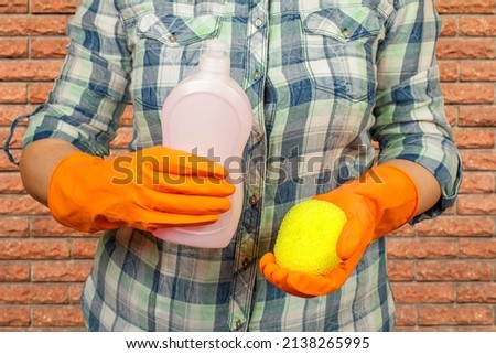 Women's hands in orange protective gloves with a bottle of dishwashing liquid and a sponge. Washing and cleaning concept.