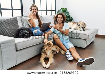 Young hispanic couple with dogs relaxing at home cheerful with a smile on face pointing with hand and finger up to the side with happy and natural expression 