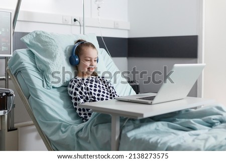 Happy sick little girl watching funny cartoons for children while resting in hospital bed. Hospitalized joyful ill little child using computer to watch funny videos on internet.