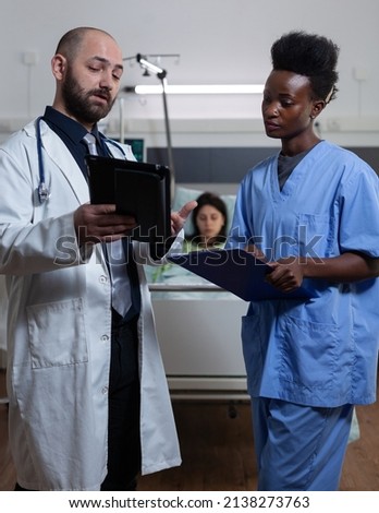 Doctor reading lab results from digital tablet to nurse holding clipboard with medical history at patient bedside in hospital ward. Health care specialists using modern technology for clinical