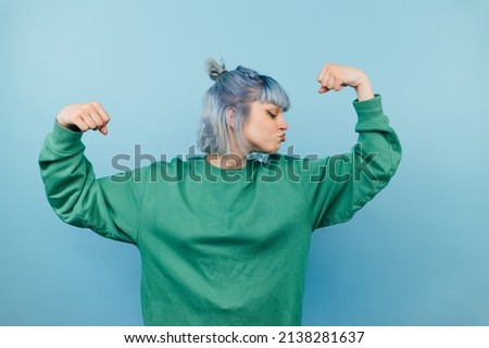 Funny hipster girl in green sweatshirt shows biceps and strength, kisses hand while standing on blue background, Royalty-Free Stock Photo #2138281637