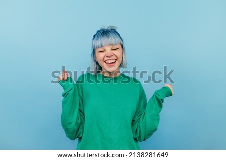 Joyful girl with blue hair in a green sweatshirt rejoices with a smile on his face and closed eyes on a blue background Royalty-Free Stock Photo #2138281649