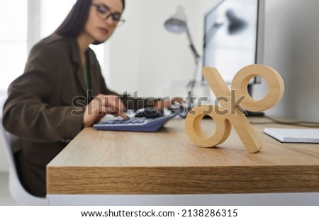 Corporate tax and interest rate. Close up of wooden percent sign standing on table on background of serious busy woman counting on calculator. Concept of interest calculation. Royalty-Free Stock Photo #2138286315