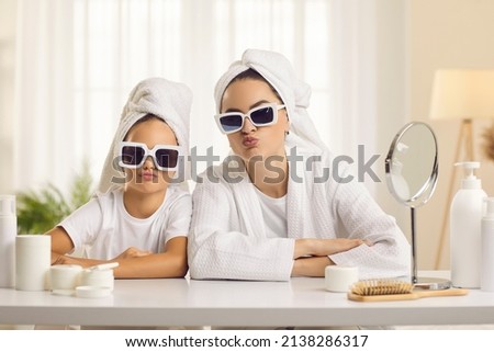 Portrait of smiling young mother and teen small daughter in towels on head and bathrobes in sunglasses send air kiss. Happy mom and little girl child enjoy home spa beauty day together.