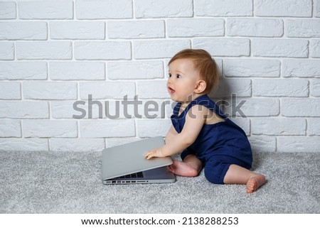 A small child 6-7 months old in a blue cotton overalls sits with an open laptop and watches educational games
