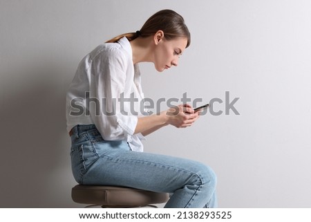 Woman with bad posture using smartphone while sitting on stool against light grey background Royalty-Free Stock Photo #2138293525