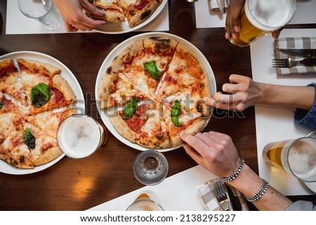 Top view restaurant table with Italian Margherita pizza and glasses of beer. Unrecognizable multiracial people's hands grabbing a sliced piece. Royalty-Free Stock Photo #2138295227