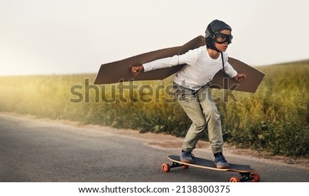 Ready, steady, fly. Shot of a young boy pretending to fly with a pair of cardboard wings while riding a skateboard outside. Royalty-Free Stock Photo #2138300375