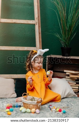 A cheerful cute girl with rabbit ears on her head in a yellow dress is decorating eggs on a green background for Easter. Funny insanely happy child is decorated. Easter for a child.
