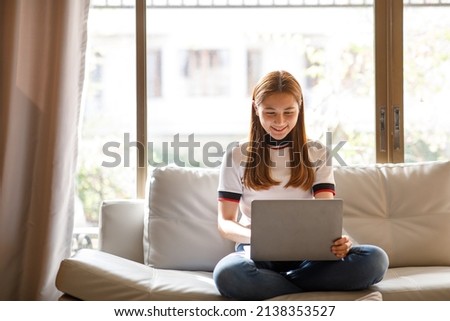 Happy smiling Asian woman sitting on couch and using laptop at living room at home, watching funny video, learning language, video calling, mother working online. High quality photo