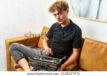 A young European guy is sitting on a sofa with a typewriter in his hands. Image in retro style. Dreamy and romantic young man relaxing in his spare time.
