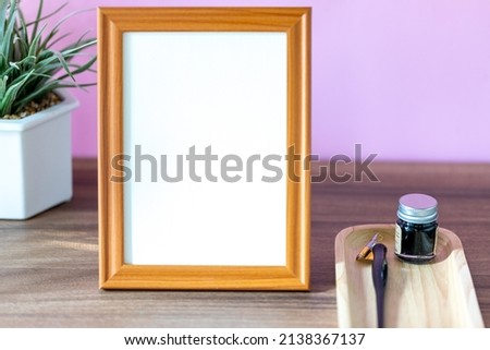 Blank vertical wooden frame beside a small houseplant and calligraphy tools, painting or artwork display mockup in front of pink wall