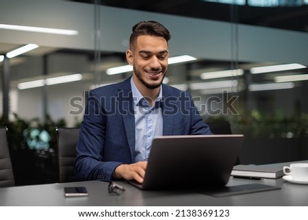 Smiling young middle eastern businessman sitting at worktable at modern office, typing on computer keyboard, sending emails to his business partners, working on marketing research, copy space Royalty-Free Stock Photo #2138369123
