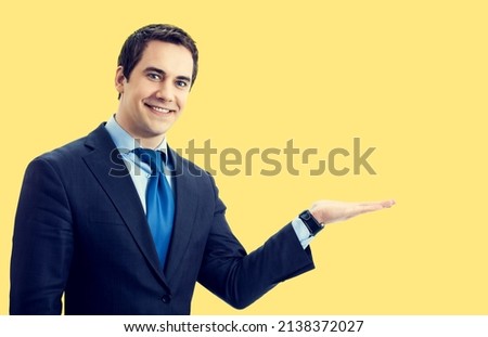 Portrait of confident businessman advertising, showing, giving or holding something, over yellow color background. Middle aged happy smiling man at studio concept image.
