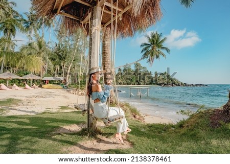 Happy Asian woman sitting on a swing on the white sand beach by the blue sea background in summer of Thailand.Travel in holidays concept.