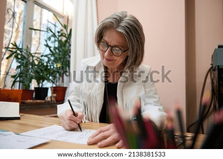 View through blurred colorful pencils holder to a fashion designer, dressmaker, mature elegant Caucasian business woman working at wooden desk, drawing sketches for clothes in her creative workshop
