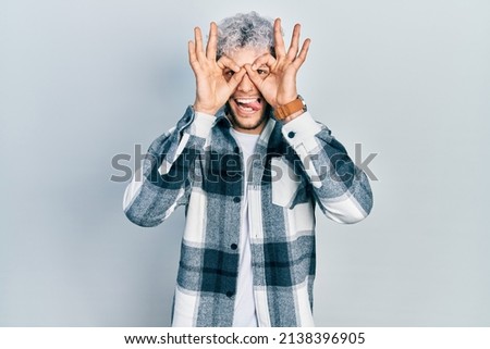 Young hispanic man with modern dyed hair wearing casual shirt doing ok gesture like binoculars sticking tongue out, eyes looking through fingers. crazy expression. 