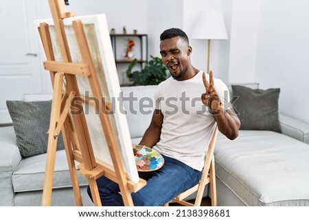 Young african man painting on canvas at home smiling with happy face winking at the camera doing victory sign with fingers. number two. 
