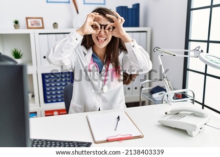 Young doctor woman wearing doctor uniform and stethoscope at the clinic doing ok gesture like binoculars sticking tongue out, eyes looking through fingers. crazy expression. 