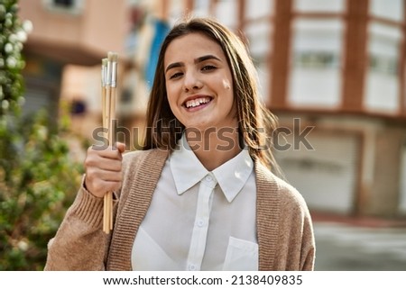 Young hispanic girl smiling happy holding paintbrushes at the city.
