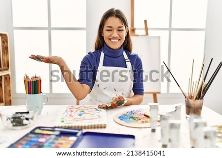 Young brunette woman at art studio with painted hands smiling cheerful presenting and pointing with palm of hand looking at the camera. 