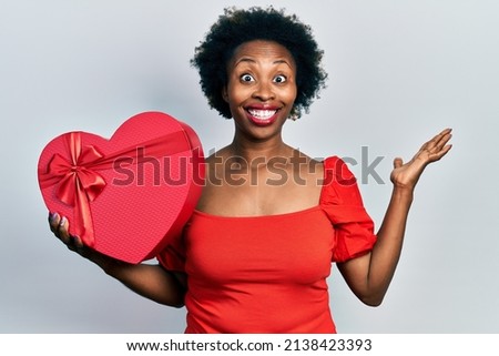 Young african american woman holding valentine gift celebrating victory with happy smile and winner expression with raised hands 