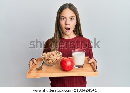 Beautiful brunette little girl holding tray with breakfast food afraid and shocked with surprise and amazed expression, fear and excited face. 