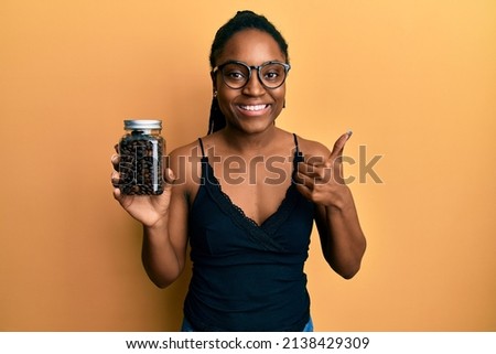 African american woman with braided hair holding jar with coffee beans smiling happy and positive, thumb up doing excellent and approval sign 