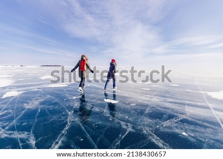 Two young women friends in red cap are skating on ice frozen of Baikal lake, winter sunny day.