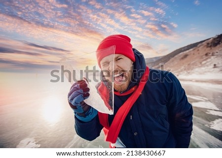 Portrait happy male tourist in red hat with shard of transparent ice in winter on Lake Baikal sunset.