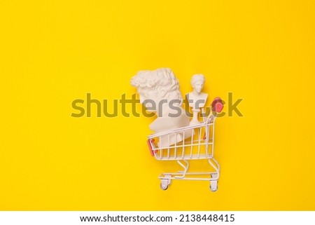 David and Venus busts in shopping trolley on yellow background. Minimal still life Royalty-Free Stock Photo #2138448415